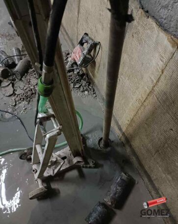 Foso industrial lleno de agua con tuberías, una escalera y escombros contra una pared de hormigón.