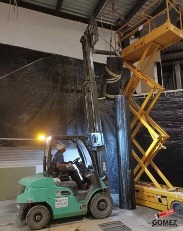 En el interior de un almacén, una carretilla elevadora verde eleva a un trabajador en una plataforma elevadora.