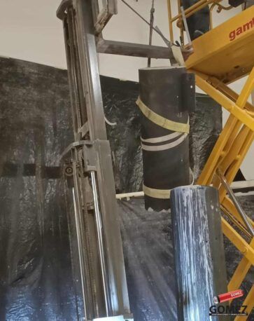 Una grúa de construcción eleva una carga cilíndrica envuelta junto a un muro de hormigón y una estructura de acero.
