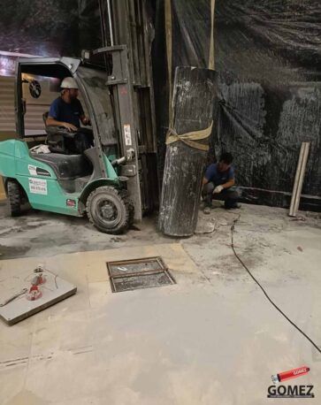 Una carretilla elevadora trabaja junto a un gran pilar industrial; un trabajador se agacha cerca de la base en una zona.