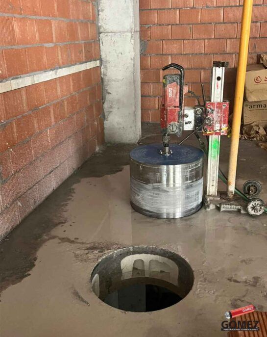 Suelo de hormigón dañado con un agujero redondo, zonas húmedas y herramientas de construcción junto a paredes de ladrillo.