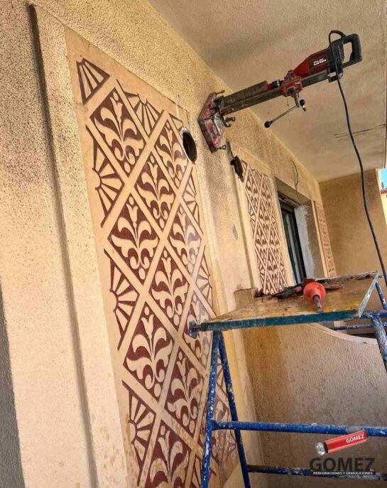 Un trabajador pinta una pared beige con paneles decorativos utilizando un andamio y una herramienta de extensión.