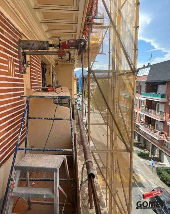 Fachada de un edificio con andamios y trabajadores en un estrecho balcón sobre una calle, envuelto en una red.