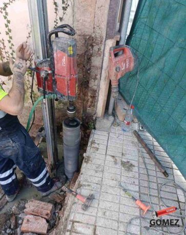 Un trabajador perfora un agujero en el hormigón junto a la pared de un edificio con una broca de núcleo rojo.