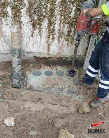 Un trabajador utiliza un taladro para cortar agujeros circulares en una losa de hormigón al aire libre, junto a una pared.