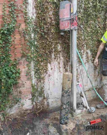 Obrero de la construcción usando un martillo neumático junto a un muro de ladrillos cubierto de enredaderas.