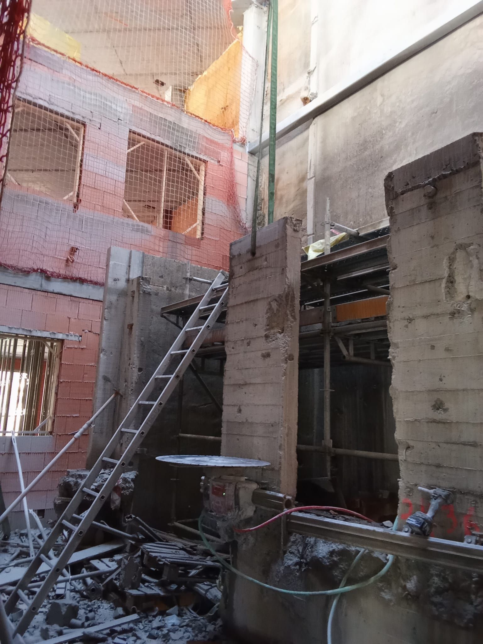 Obra en construcción con muros de hormigón sin terminar, una escalera, andamios y escombros en un patio estrecho.