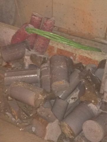 Tuberías grises rotas y escombros amontonados en un rincón sucio, con mangueras verdes y rojas pegadas a una pared manchada.