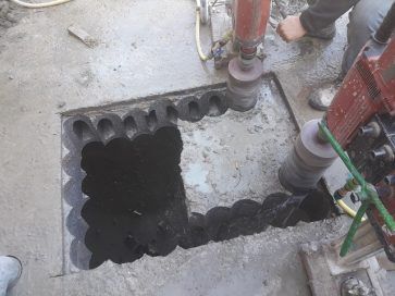 Un hueco cuadrado en un suelo de hormigón alrededor de tuberías y un taladro, con escombros y herramientas cerca.