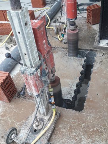 Perforadora de construcción taladrando en el piso de concreto junto a una columna de soporte roja.