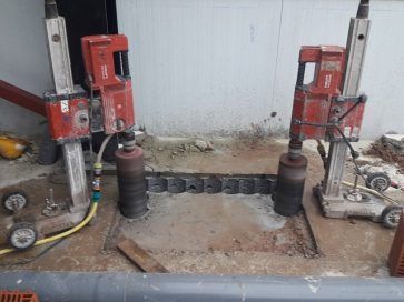 Dos perforadoras rojas taladrando hormigón junto a un muro, con polvo y escombros en el suelo.