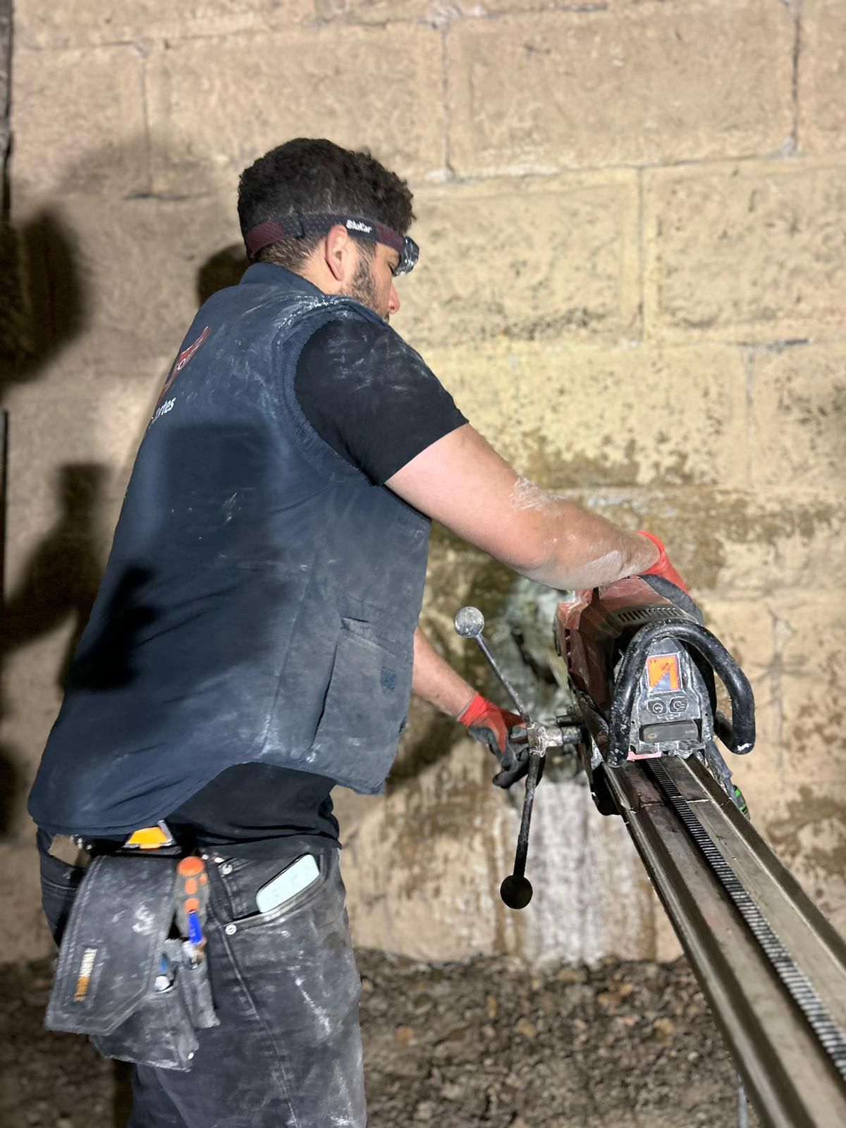 Un trabajador, vestido con ropa oscura y gafas de seguridad, corta un muro de hormigón con una sierra eléctrica, con las herramientas cubiertas de polvo.