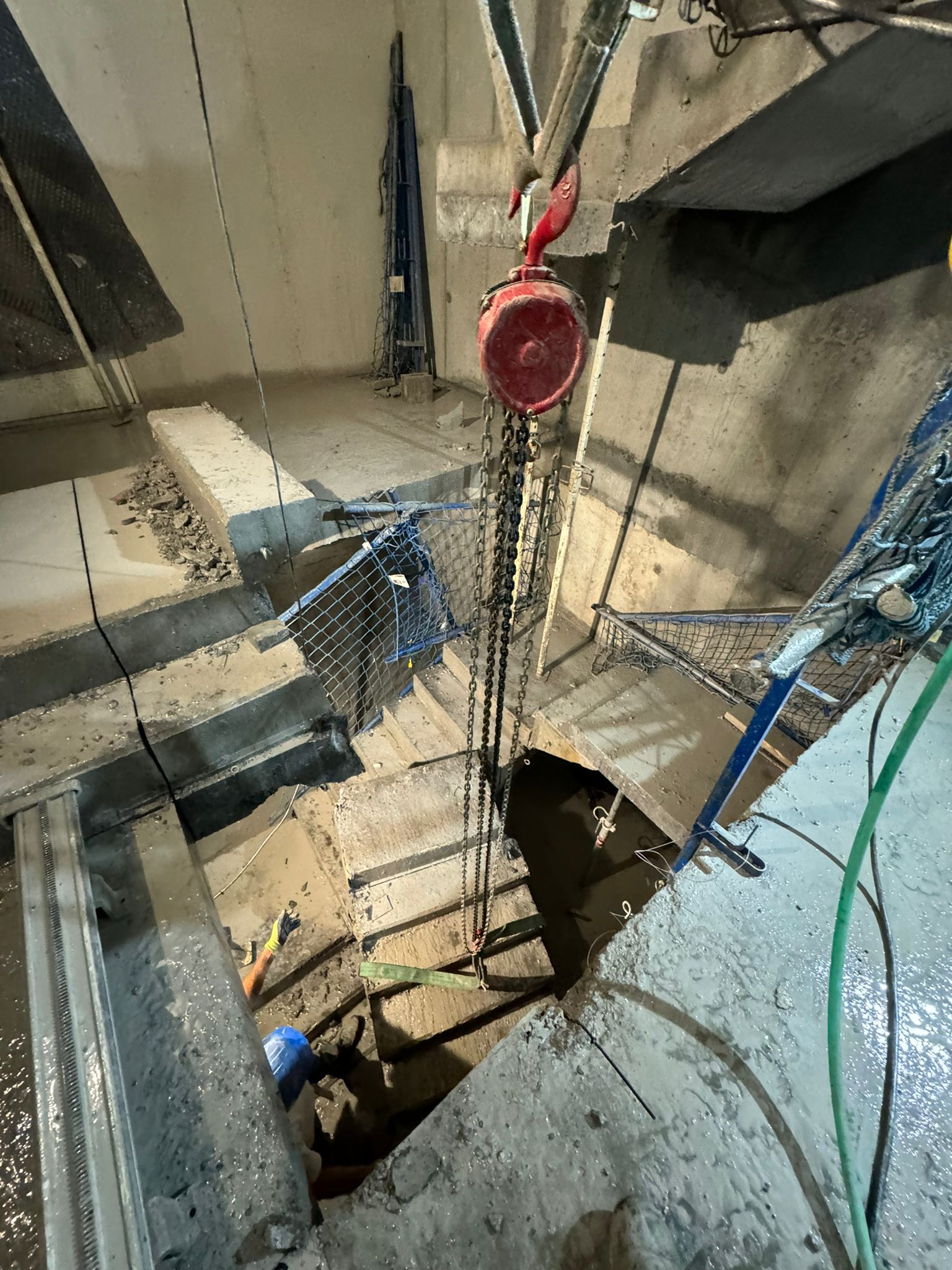 Pozo de construcción con polea roja y cuerdas colgando sobre una abertura en el suelo de hormigón.