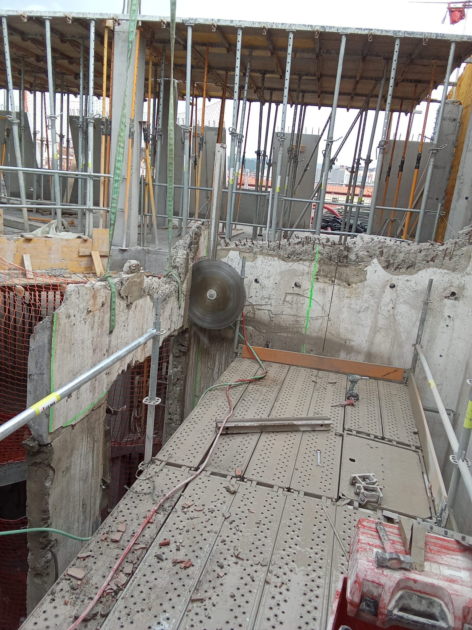 Obra en construcción con columnas de barras de refuerzo, muros de hormigón y una estrecha pasarela de madera sobre una zanja.