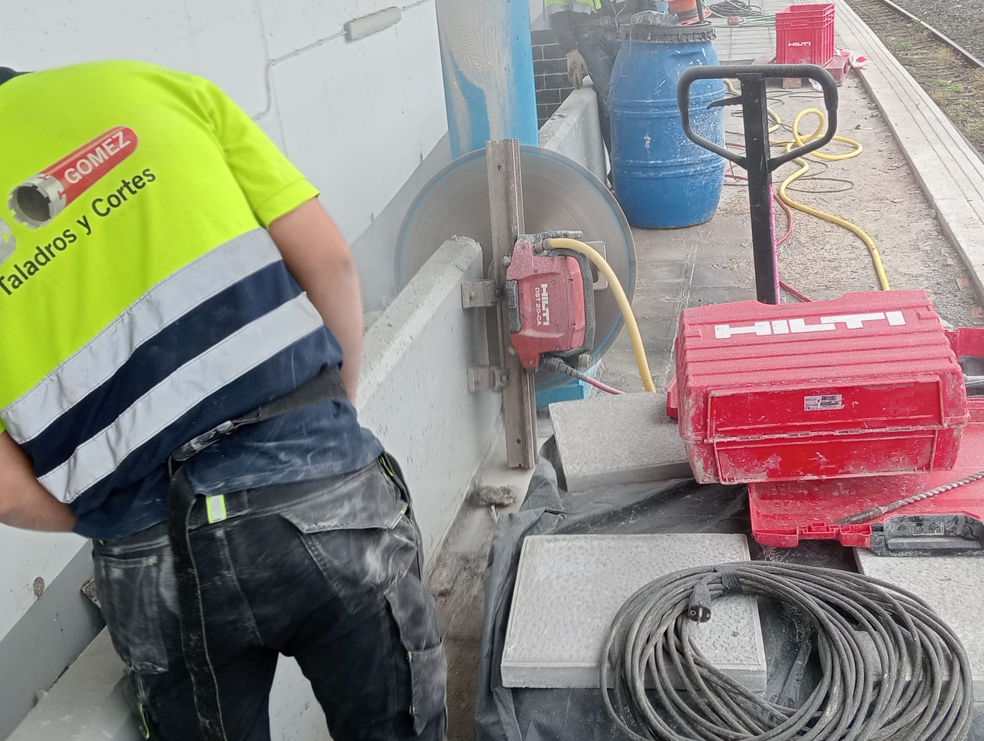 Obrero cortando hormigón con una sierra en una zona de construcción, con herramientas y cables cerca.