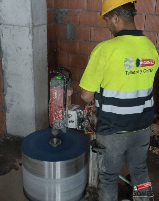 Un trabajador opera una perforadora de hormigón en una gran losa gris en el interior de un edificio; lleva un casco.