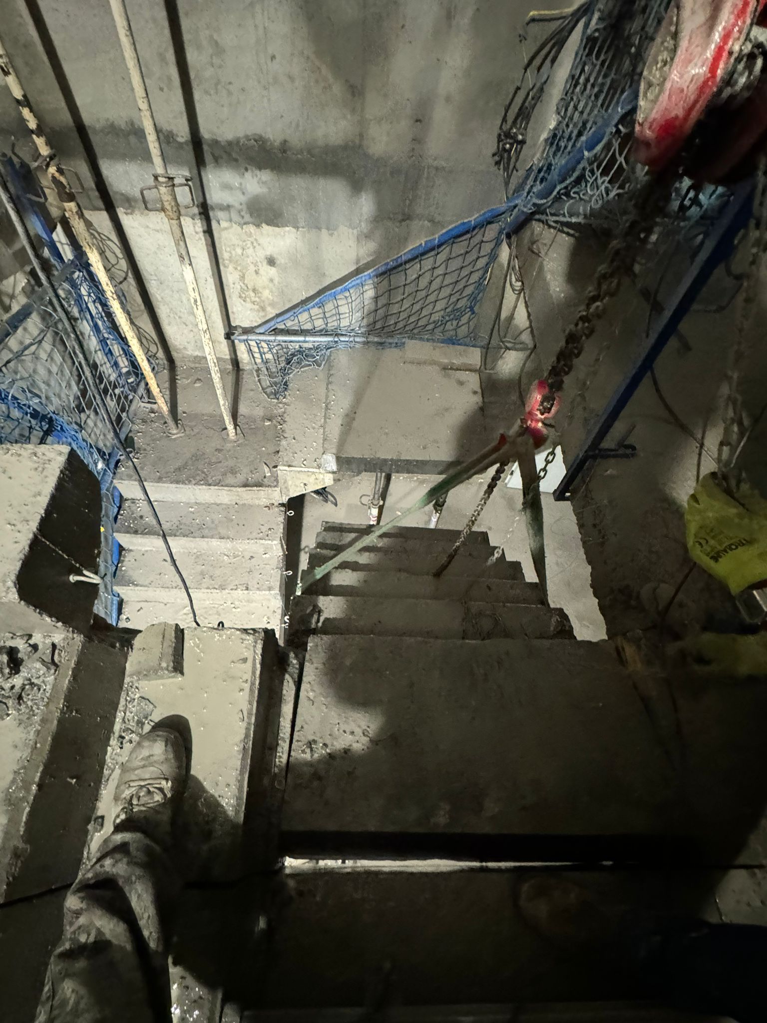 Escalera de hormigón oscuro con cuerdas azules y barandillas de metal, vista desde arriba bajo una iluminación intensa.
