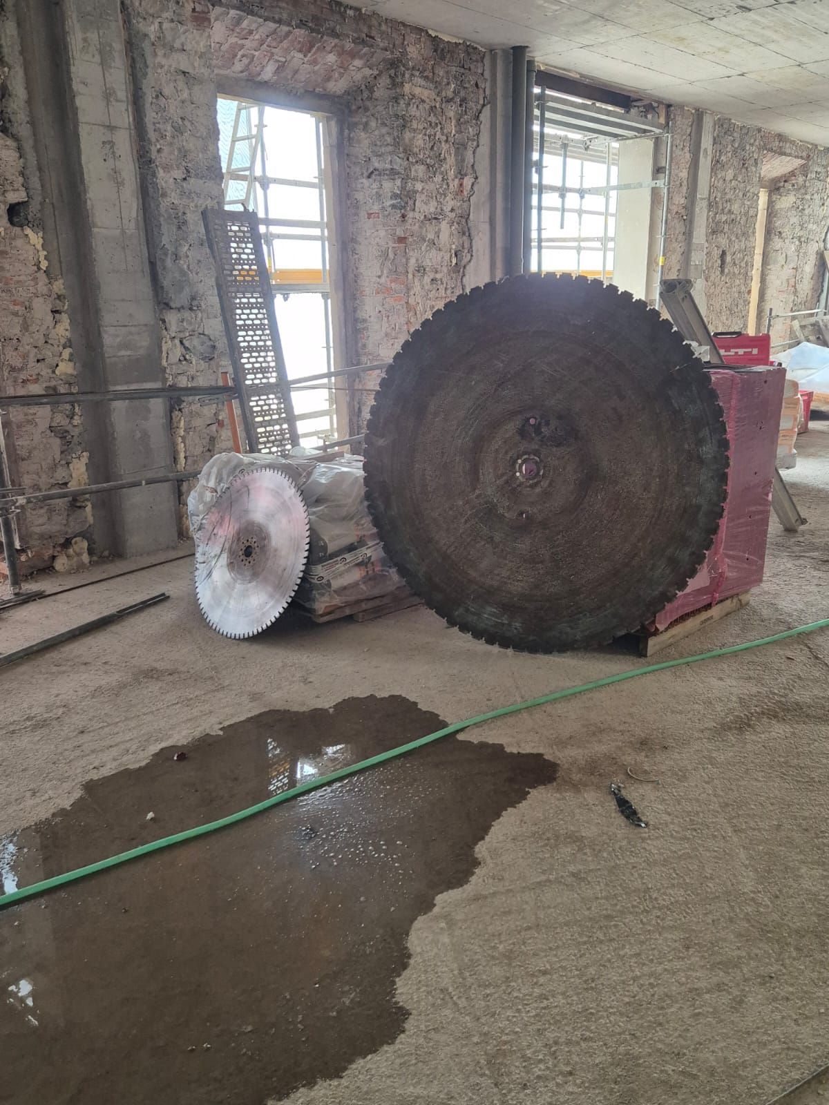 Grandes hojas de sierra y troncos en el suelo de un taller, con un charco y una manguera verde cerca de las ventanas.