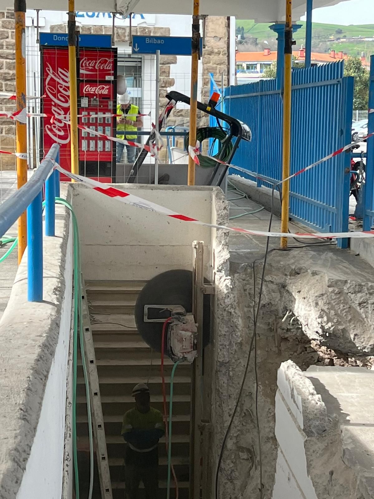 Zona de construcción con una escalera abierta y una entrada tipo túnel, acordonadas con cinta y vallas azules.