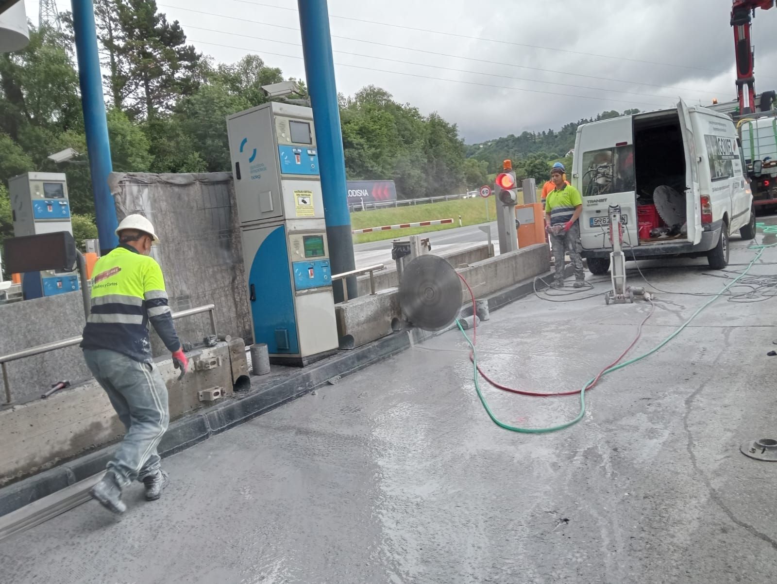 Obreros pavimentando una carretera mojada junto a una furgoneta blanca y maquinaria azul bajo un cielo nublado.