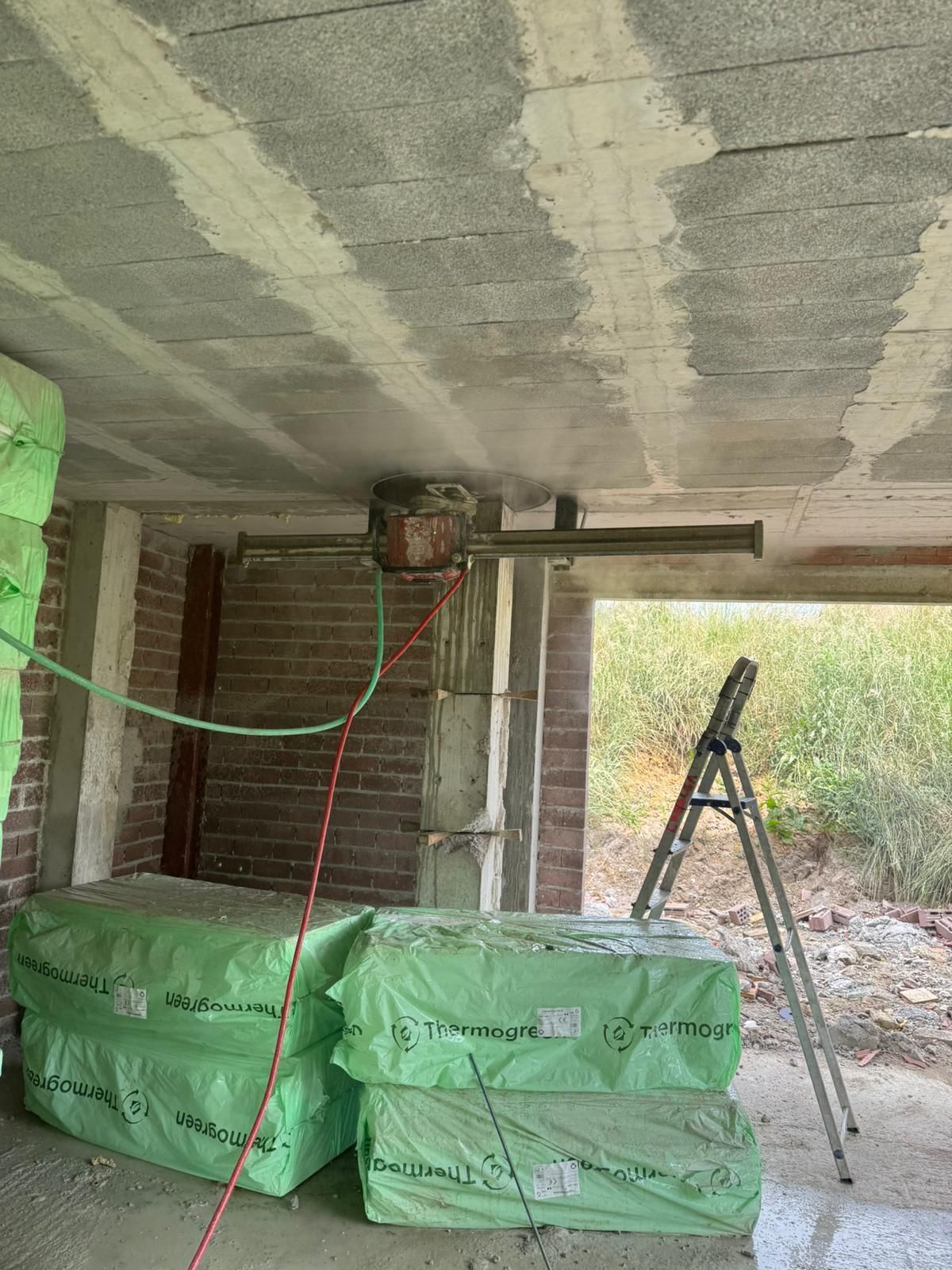 Obra en construcción con sacos verdes apilados, escalera, techo de hormigón visto y una herramienta eléctrica colgando.