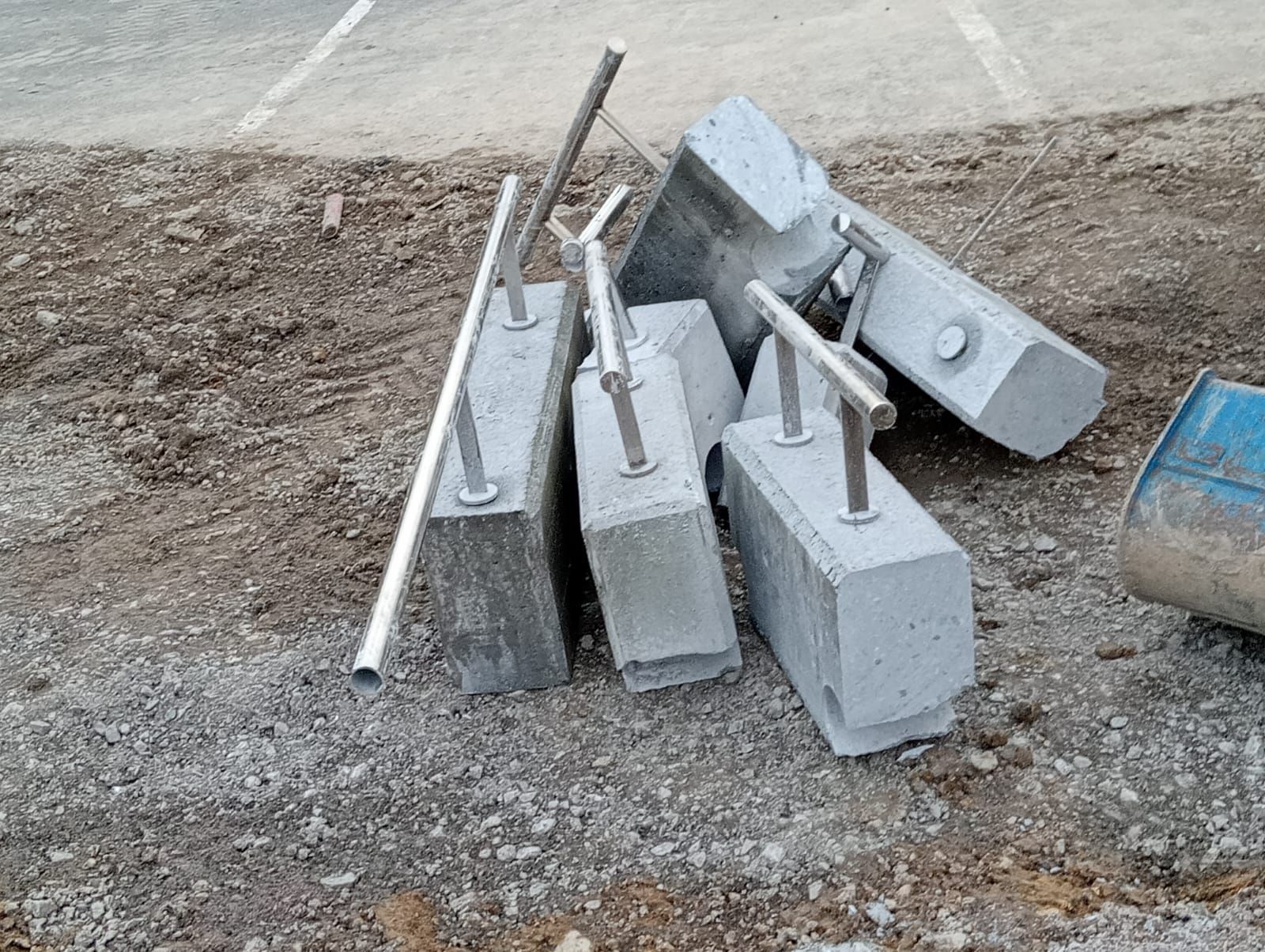 Montón de barreras de hormigón gris y postes metálicos sobre grava junto a una carretera, con un barril azul a la derecha.
