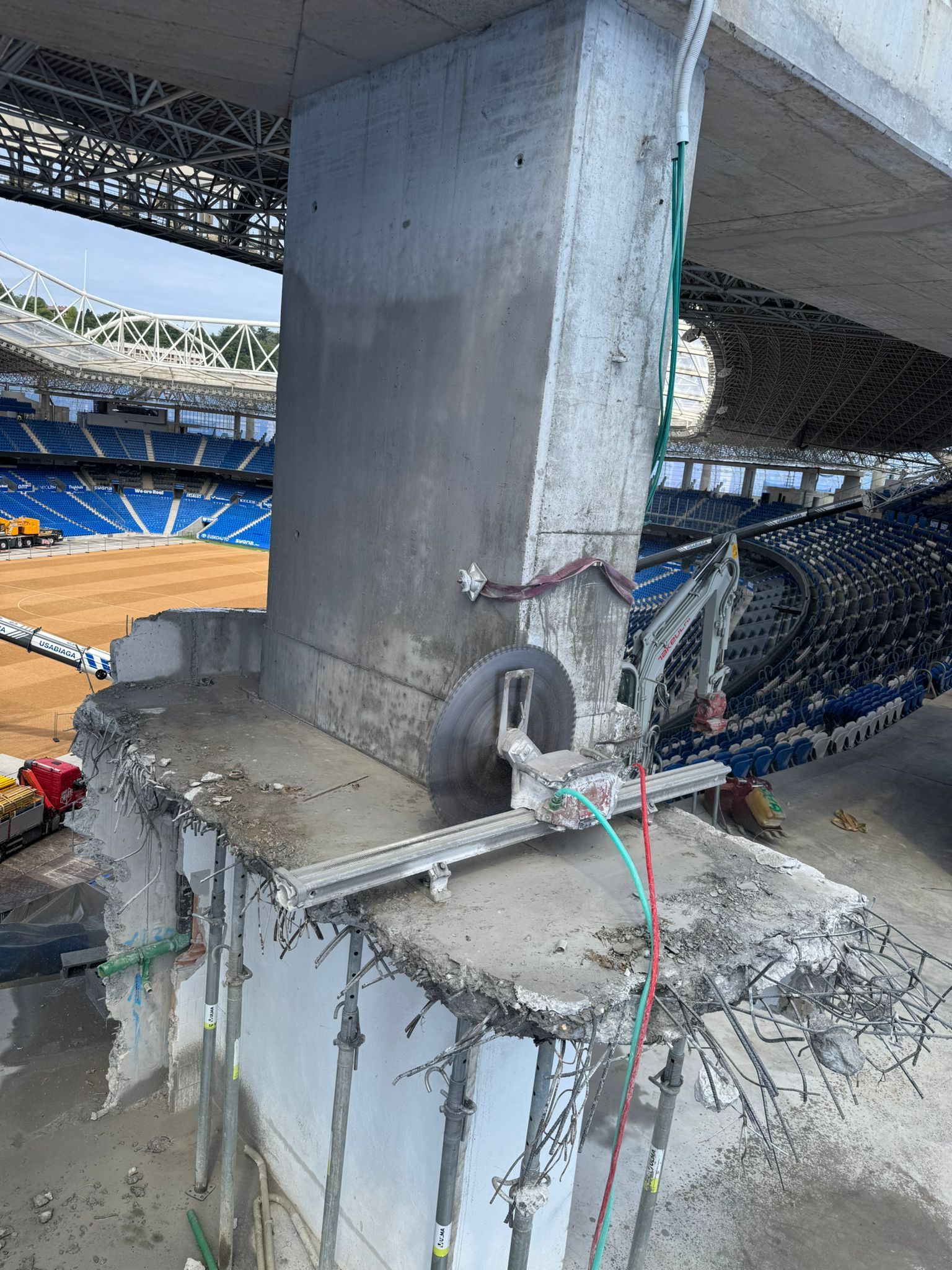 Columna de soporte de hormigón dentro de un estadio, con cables de construcción y asientos azules al fondo.