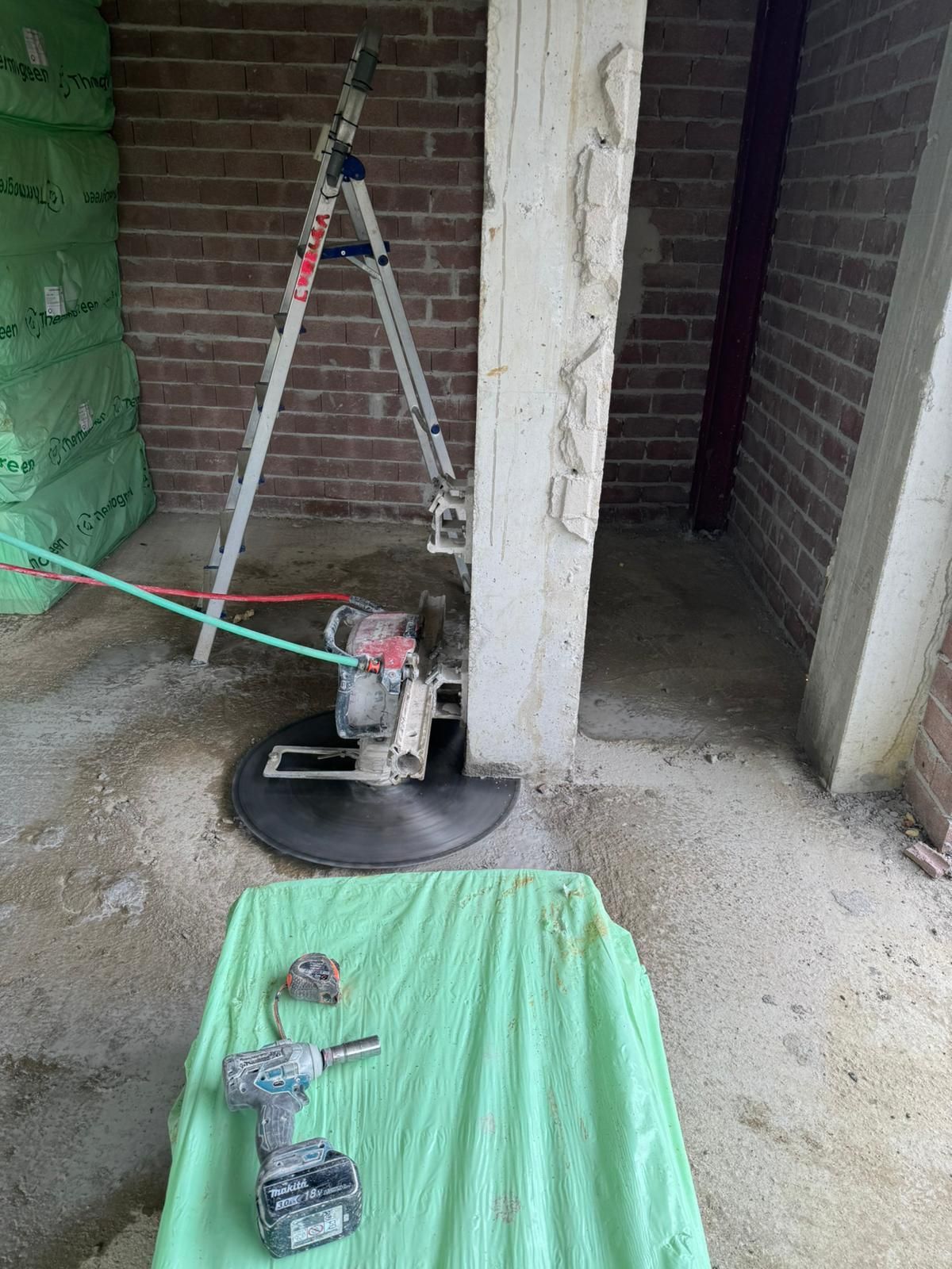 Obra en construcción con una escalera, una columna de hormigón, una herramienta eléctrica y una lona verde en el suelo.