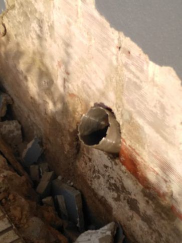 Pared dañada con un agujero circular que deja al descubierto escombros y un interior oscuro, con manchas rojizas cerca.