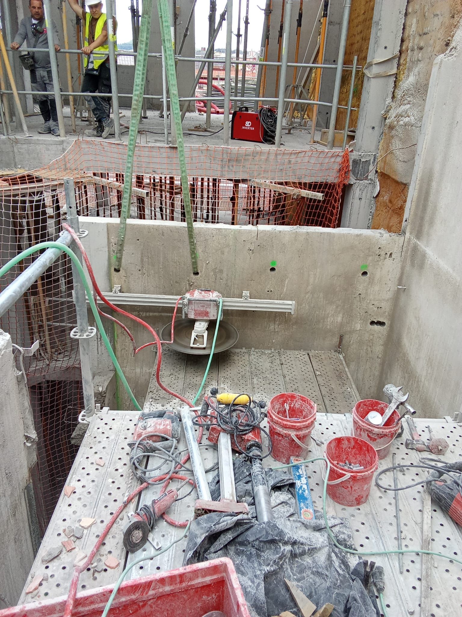 Obra en construcción con barras de refuerzo, herramientas, cubos rojos y muros de hormigón en un foso estrecho.