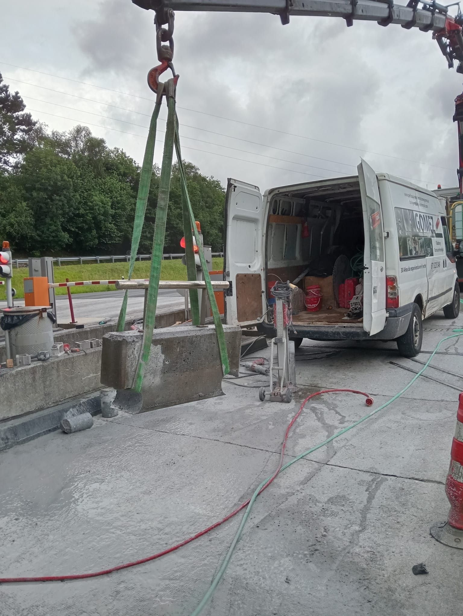 Obra en construcción con una grúa levantando un bloque de hormigón junto a una furgoneta de obra abierta en una carretera.
