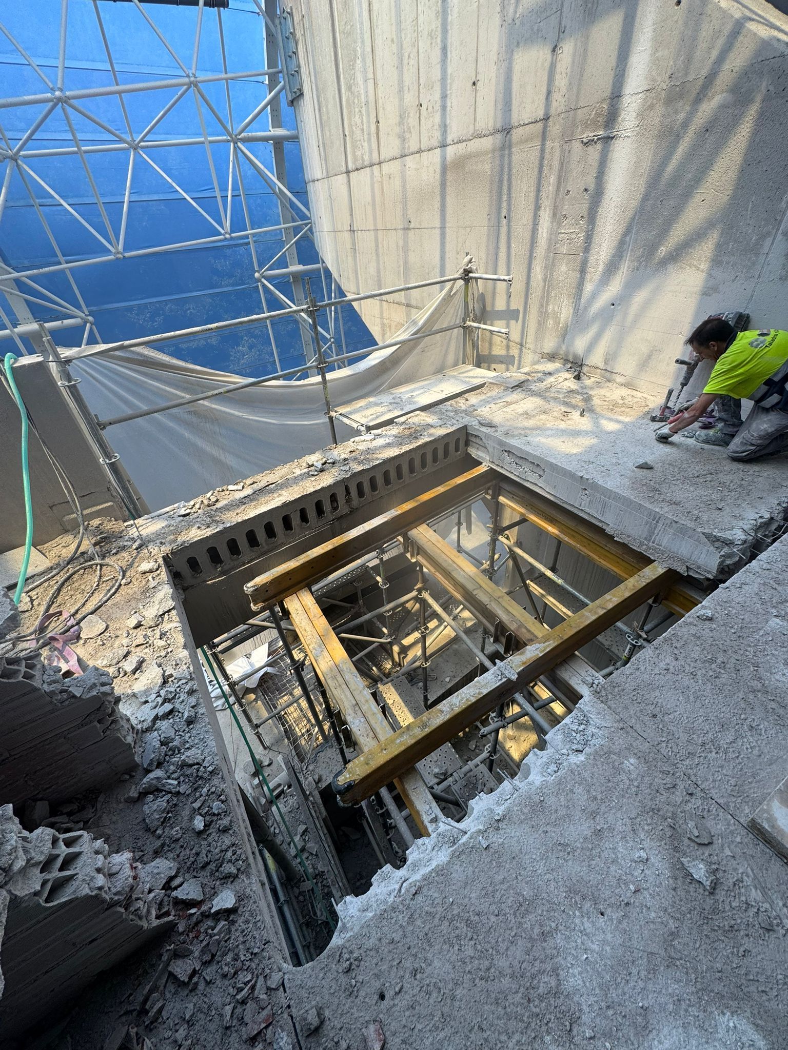 Obreros de la construcción junto a una abertura cubierta con andamios en un piso de concreto, con escombros y herramientas a su alrededor.
