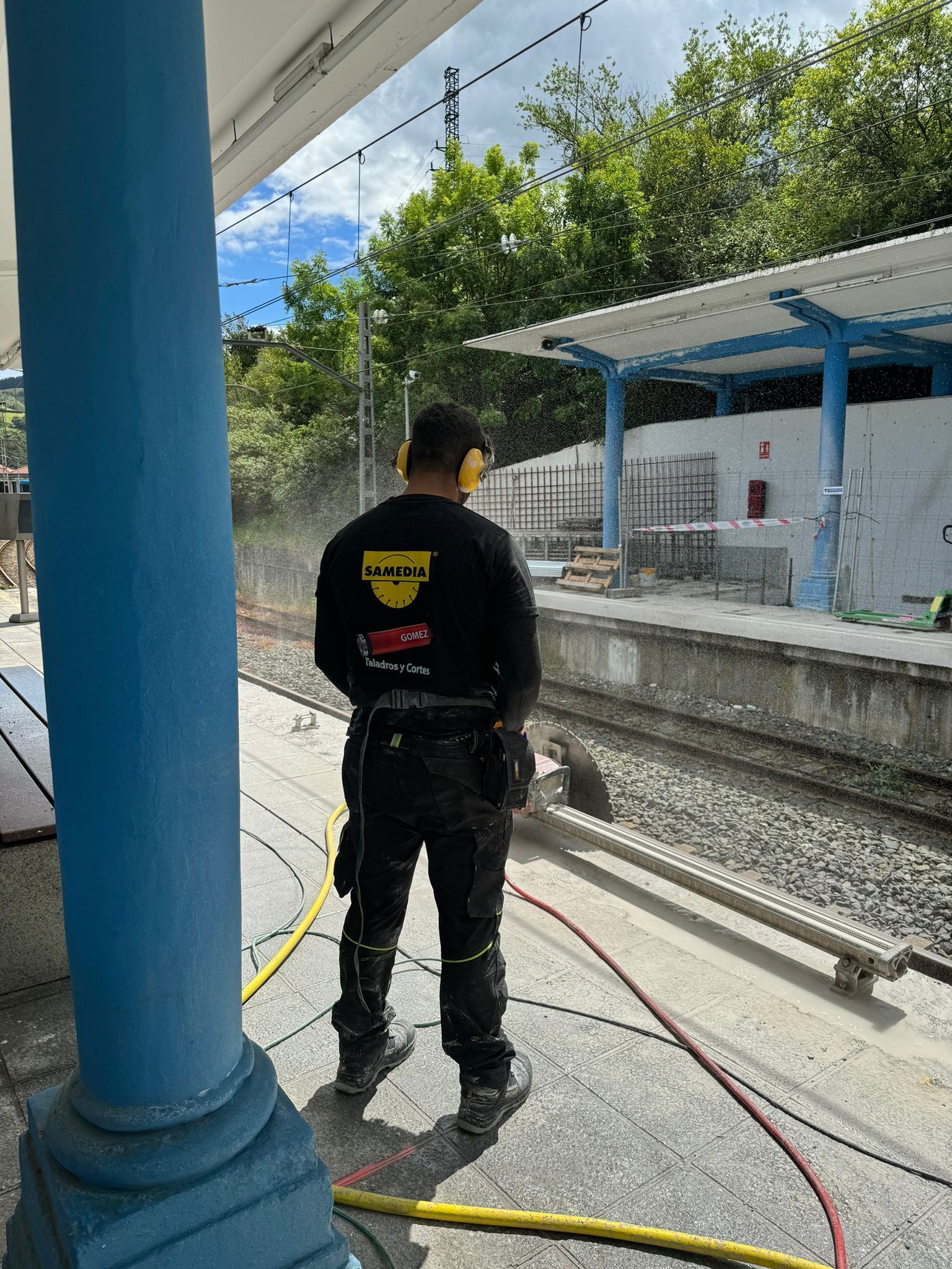 Un trabajador vestido de negro se encuentra en un andén de ferrocarril junto a columnas azules y una vía férrea, sujetando un cable.