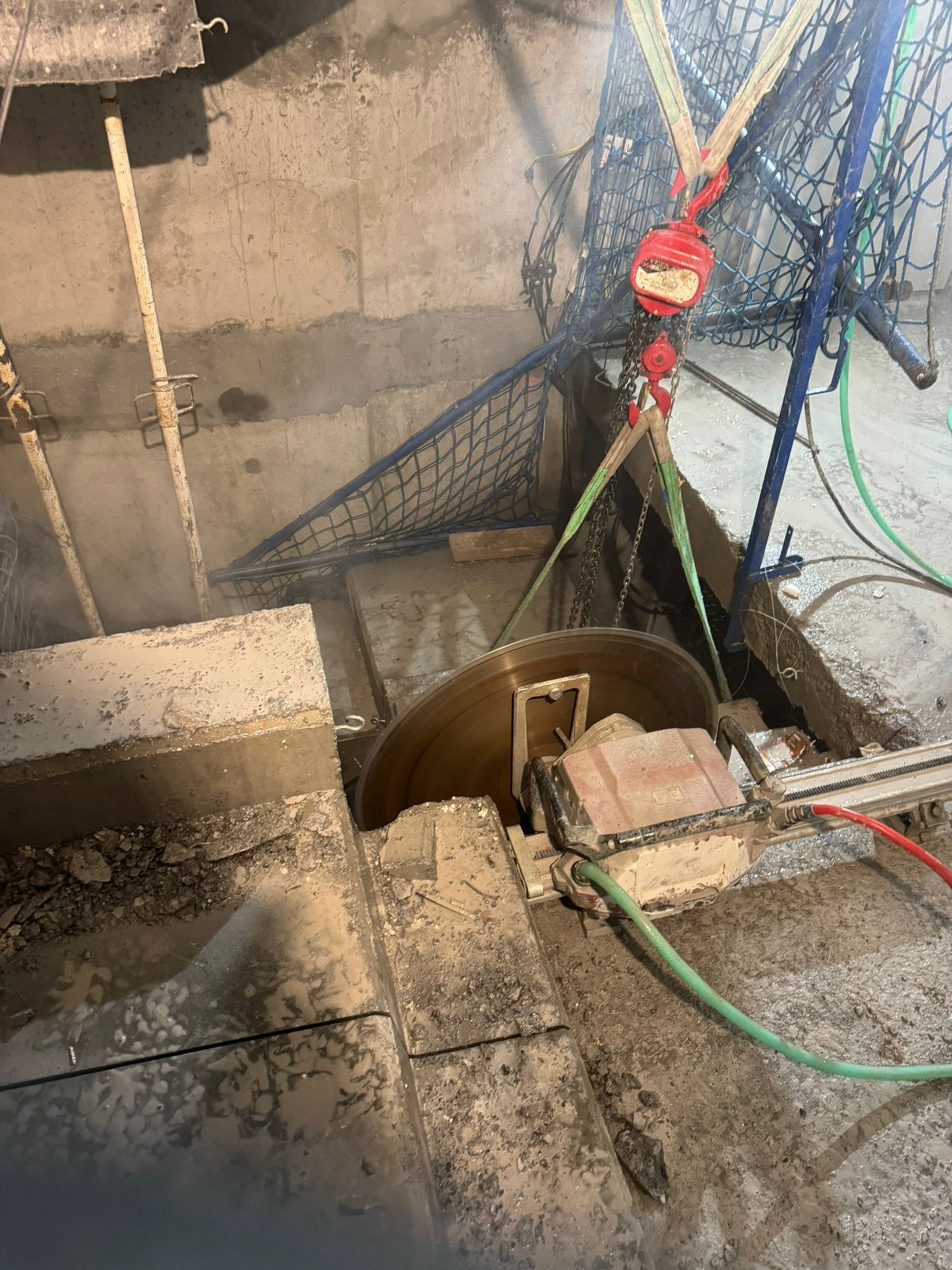 Escalera de hormigón con un cubo cubierto, una manguera verde y una válvula roja junto a una pared y un suelo húmedos.
