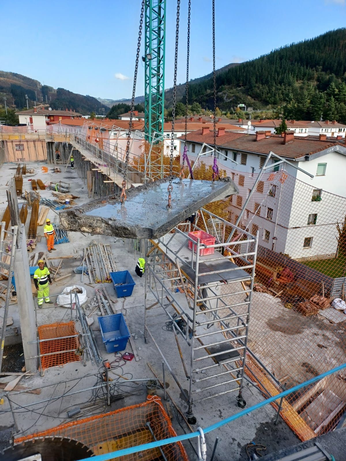 Obra en construcción con trabajadores, andamios, grúa y vertido de hormigón junto a edificios en la ladera.