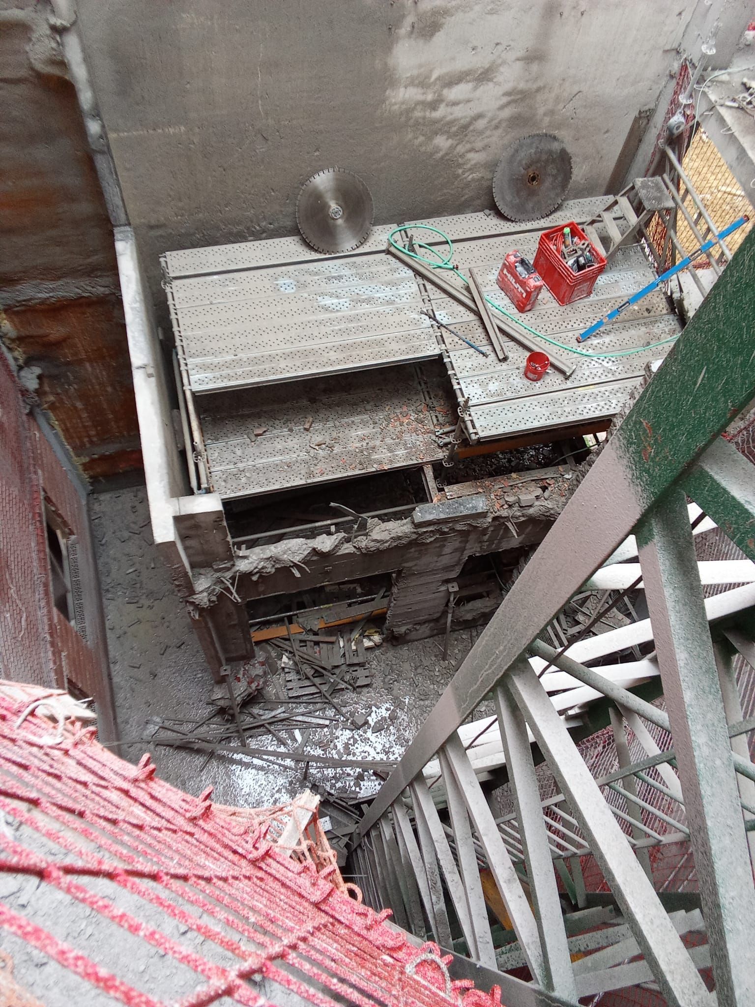 Vista hacia abajo de una escalera y una plataforma de construcción con vigas metálicas, una red roja y escombros.