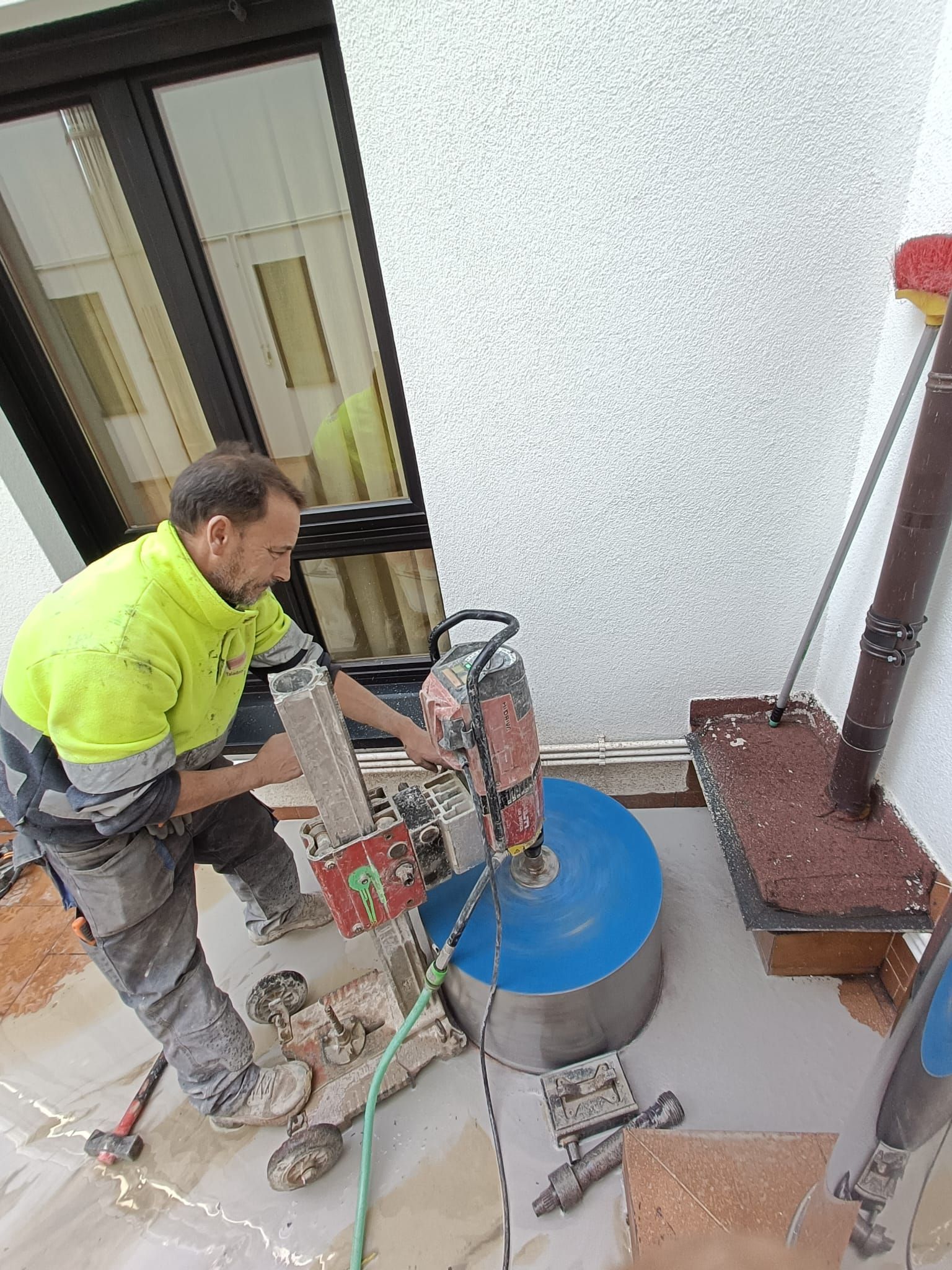 Un trabajador utiliza una perforadora de núcleos en una losa de hormigón junto a un edificio, con agua azul que recoge el polvo.