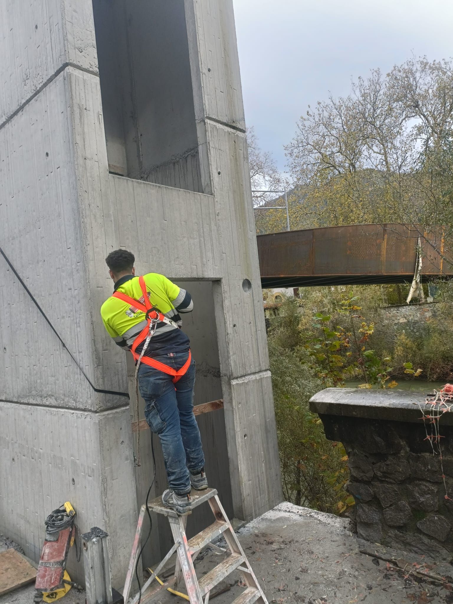 Un trabajador con un chaleco de seguridad de color neón está subido a una escalera, inspeccionando la pared de hormigón de un edificio.