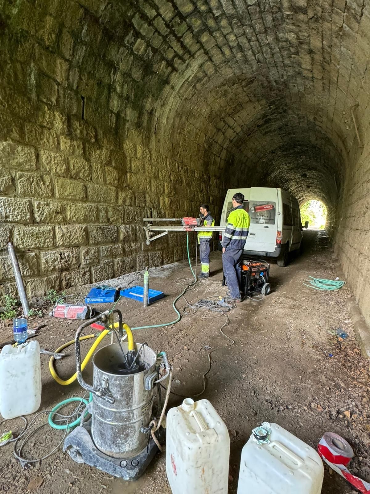 Trabajadores y equipos dentro de un túnel de piedra, con herramientas y un camión utilitario blanco al fondo.