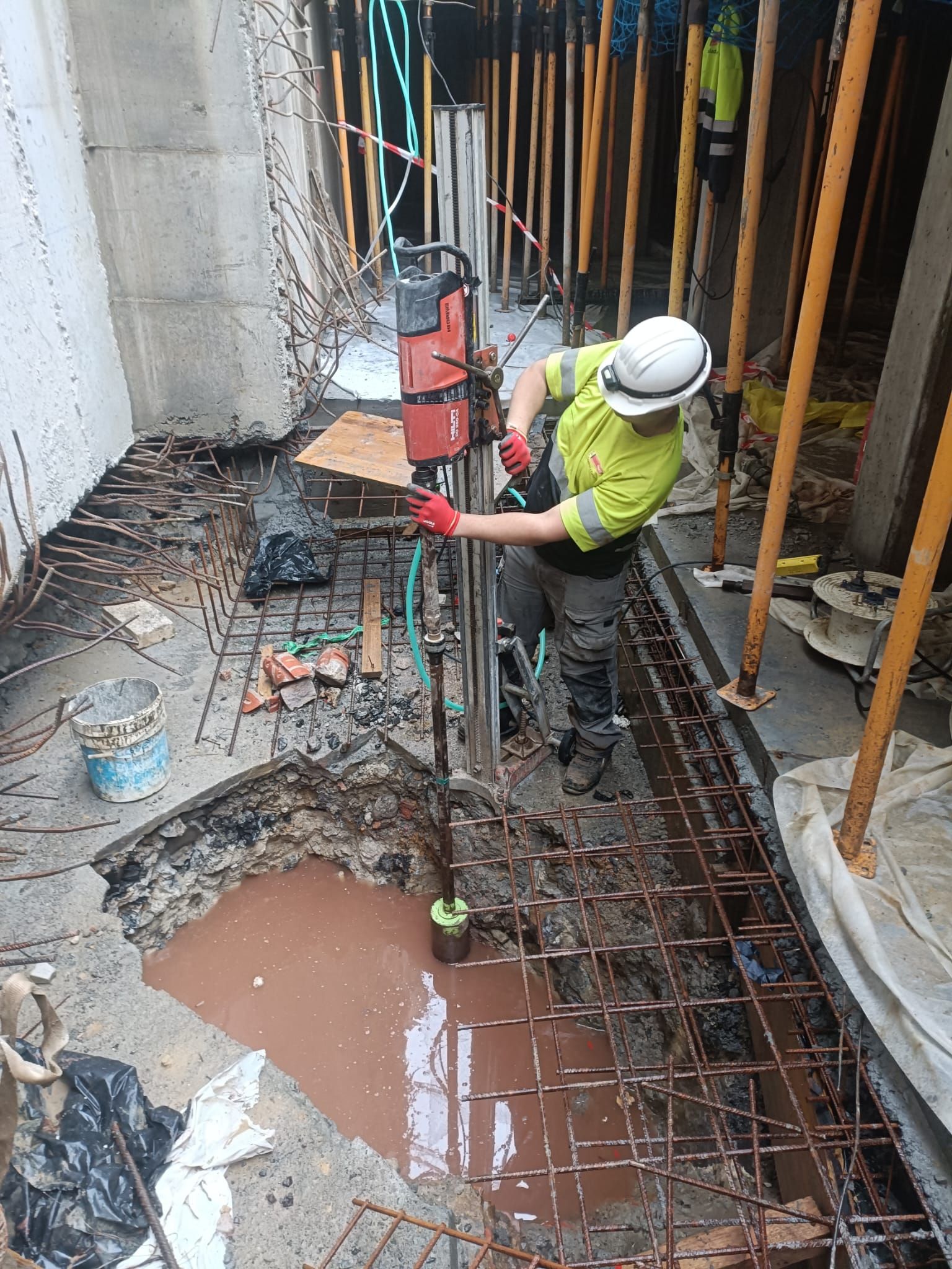 Un trabajador con equipo de seguridad perfora una zanja de construcción con barras de refuerzo y agua fangosa cerca.
