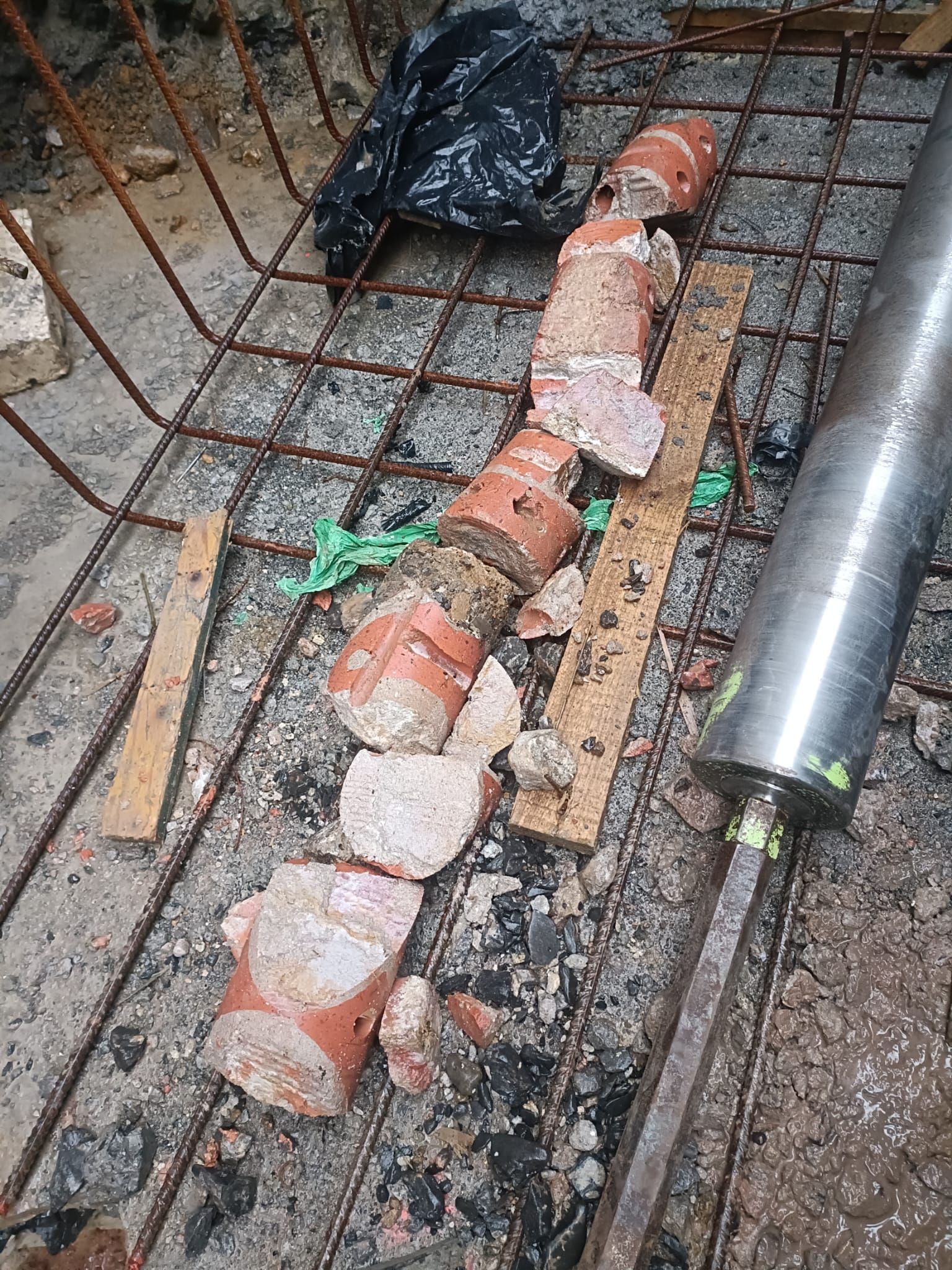 Sección rota de tubería naranja en una zanja de construcción húmeda junto a barras de refuerzo y herramientas.