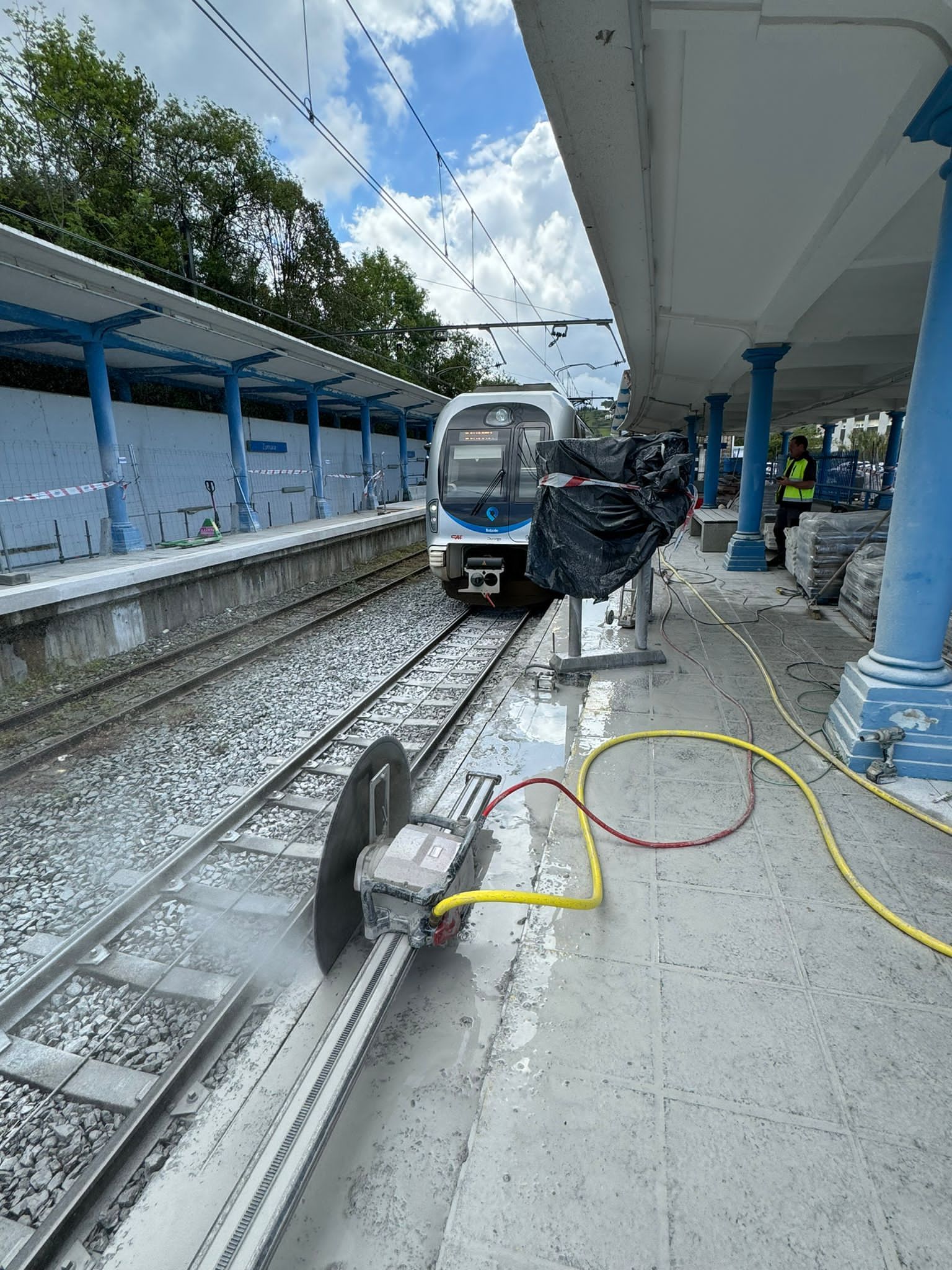 Tren en el andén de la estación con trabajadores y mangueras junto a las vías bajo un toldo azul.