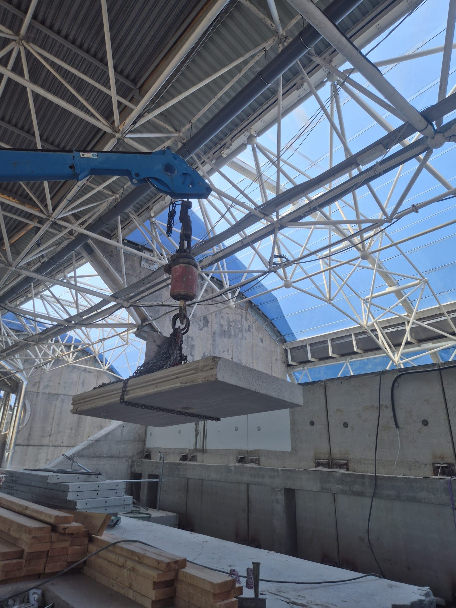 Grúa elevando una losa de hormigón en el interior de una obra en construcción con cerchas de acero a la vista.