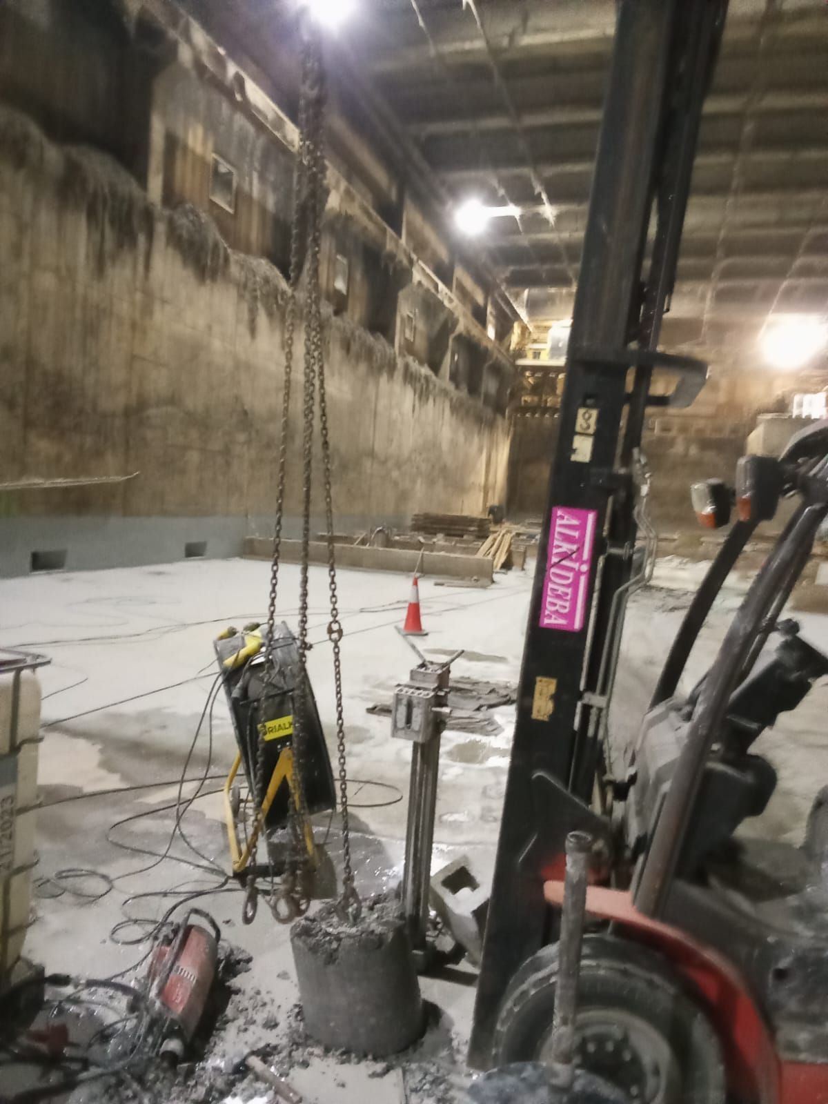 Interior de una obra en construcción con excavadora, suelo de hormigón, cadenas y cables colgantes bajo iluminación industrial.