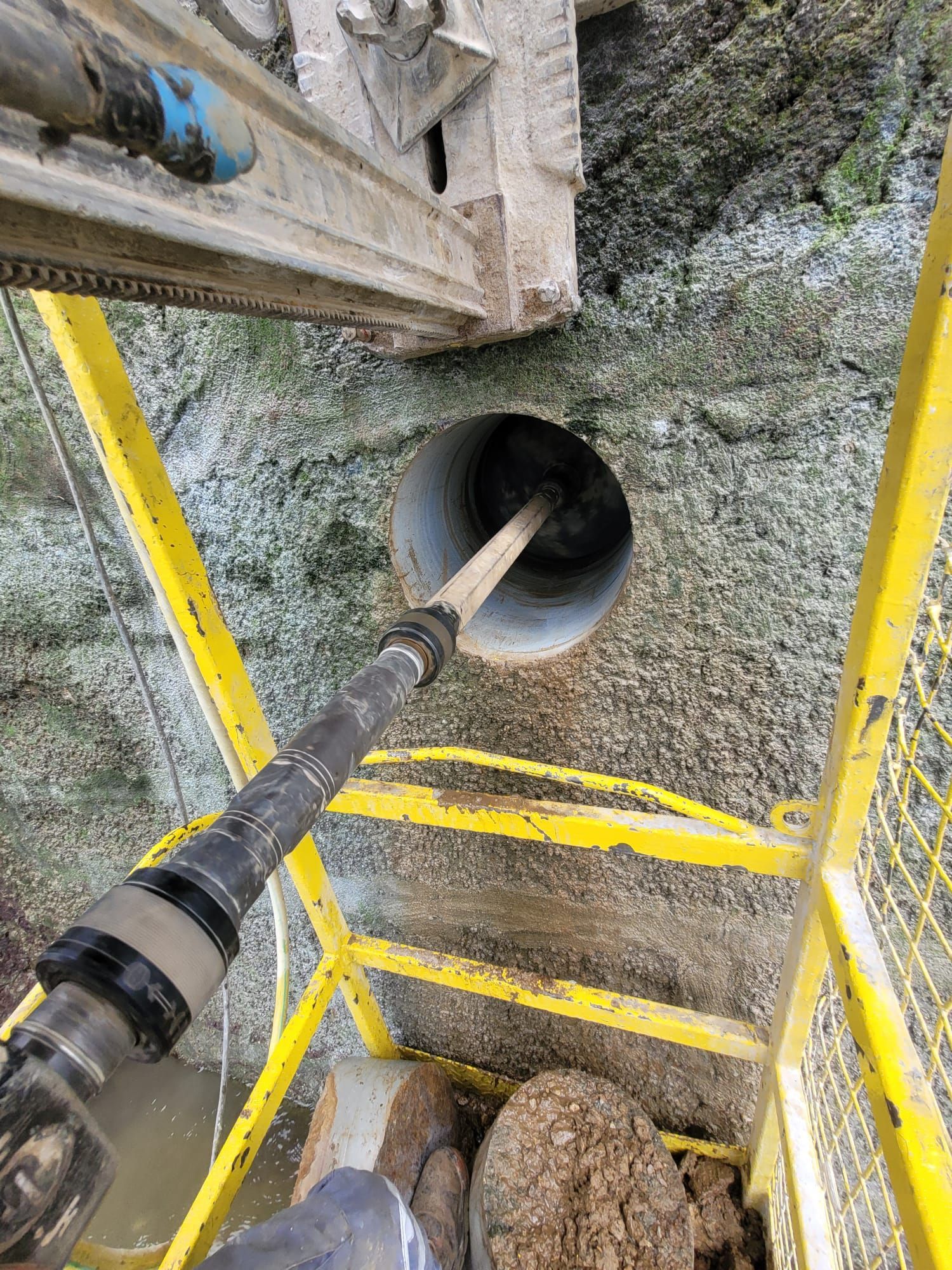 Un trabajador baja una manguera a una alcantarilla redonda junto a barandillas de seguridad amarillas en una obra rocosa.