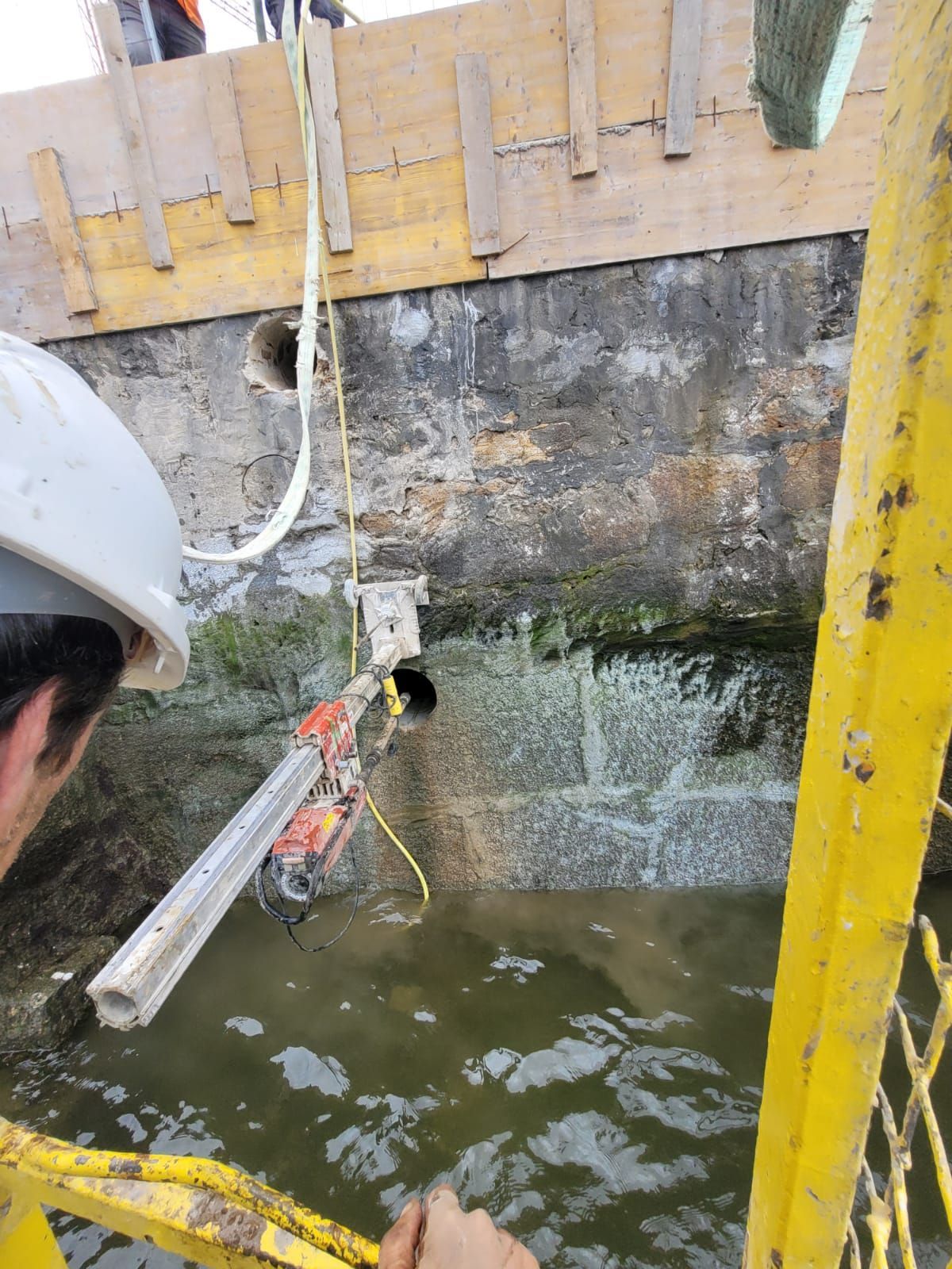 Obrero de la construcción con casco utilizando una manguera para rociar agua contra un muro de hormigón en una zona inundada.