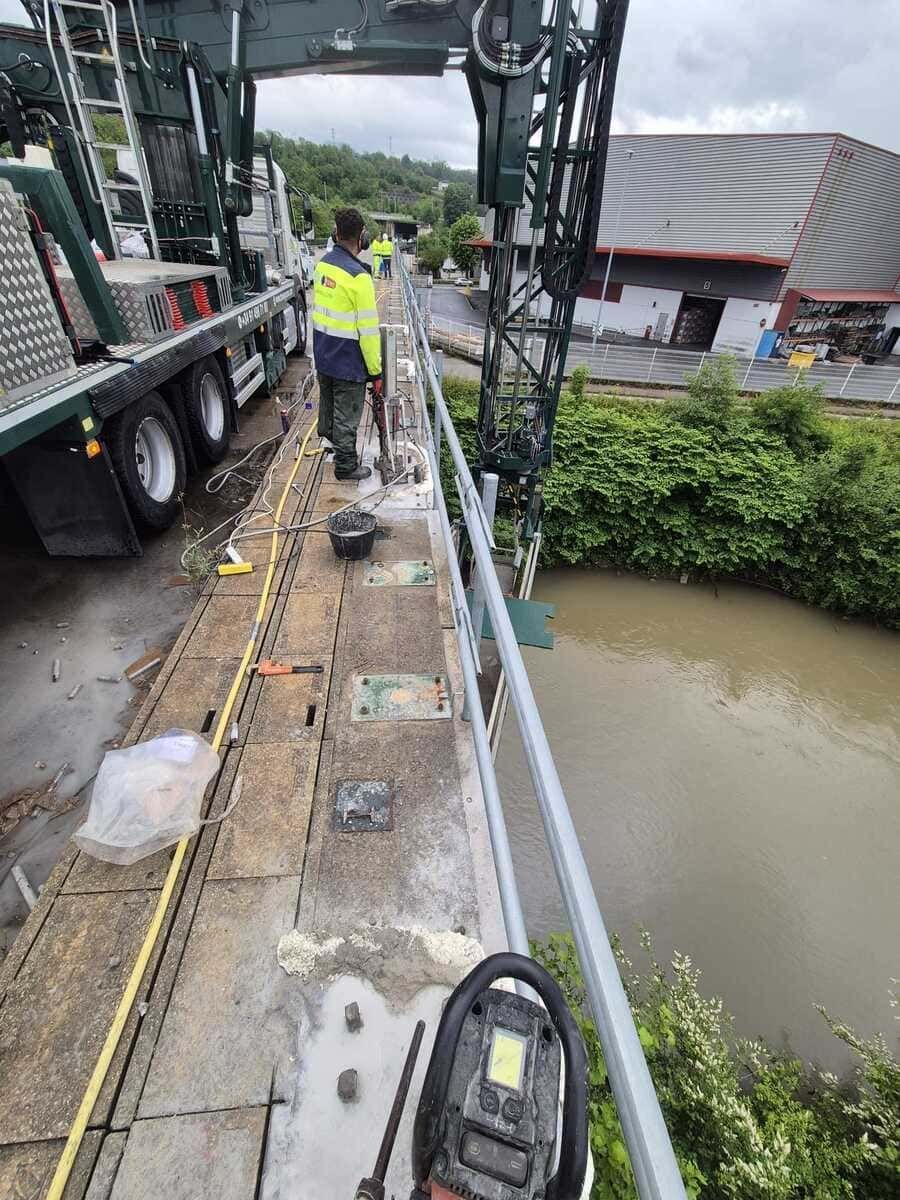 Un trabajador con un chaleco reflectante se encuentra en un puente estrecho junto a un camión, con vistas a un río lodoso que discurre abajo.