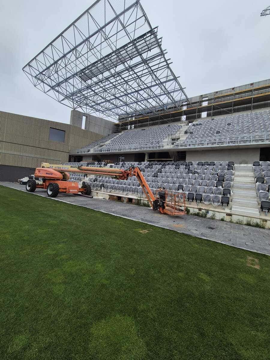 Maquinaria de construcción en el campo de un estadio, bajo una tribuna parcialmente cubierta, con césped verde en primer plano.