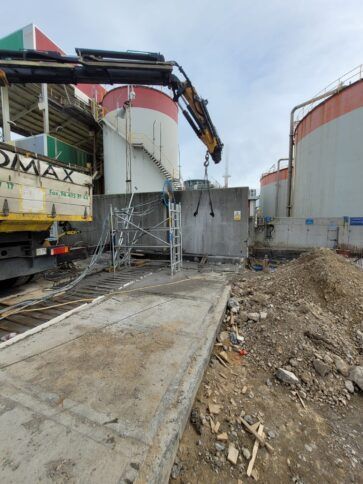 Obra en construcción con una grúa levantando un panel junto a grandes tanques industriales y un camino de hormigón.