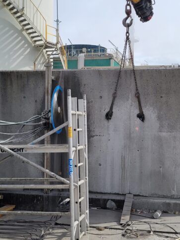 Obra en construcción con un muro de hormigón, cadenas de polipasto suspendidas y una escalera apoyada cerca.